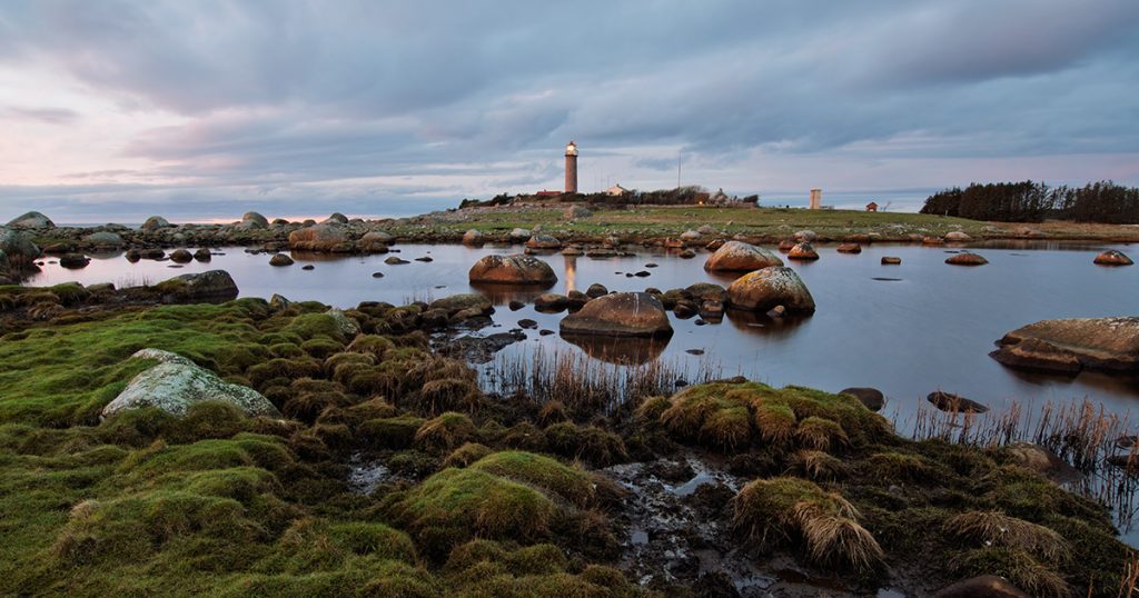 Lista fyr i skumring med lys i lykta, våtmark og steiner i forgrunnen, og stille vann som speiler himmelen.