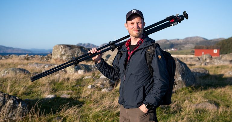 Ivan Mæland med kamerastativ over skulderen i kystlandskap